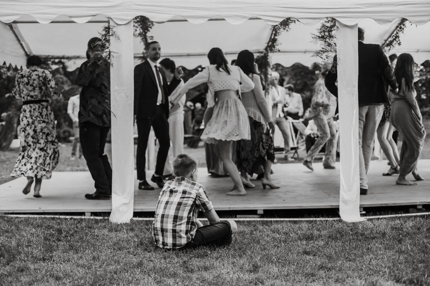 A boy sitting alone on the grass, watching guests dance under a marquee, black and white