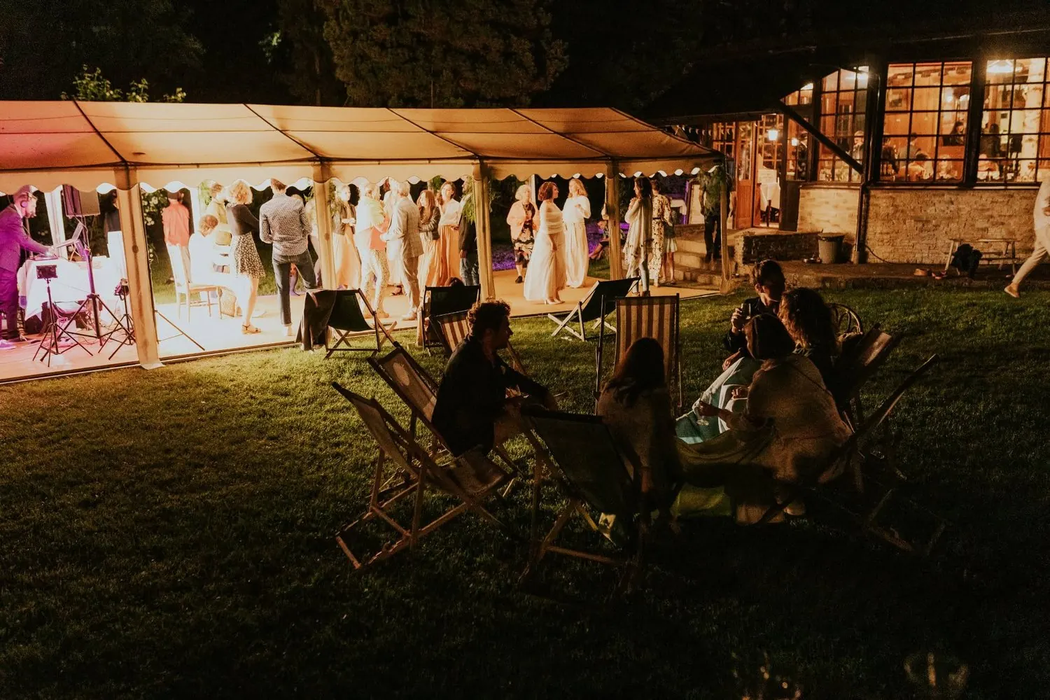 A lit marquee at night: the DJ on the left, guests resting in deckchairs on the dark lawn